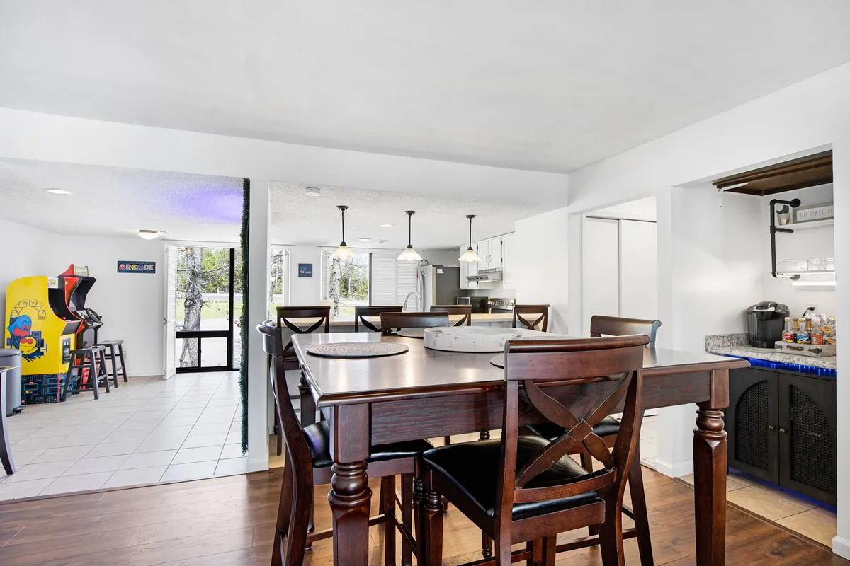 A lively view of an open dining and lounge area featuring a dining table adjacent to an arcade game, with the kitchen area in the background. The scene conveys casual gatherings, game nights, and multifunctional spaces for entertainment.