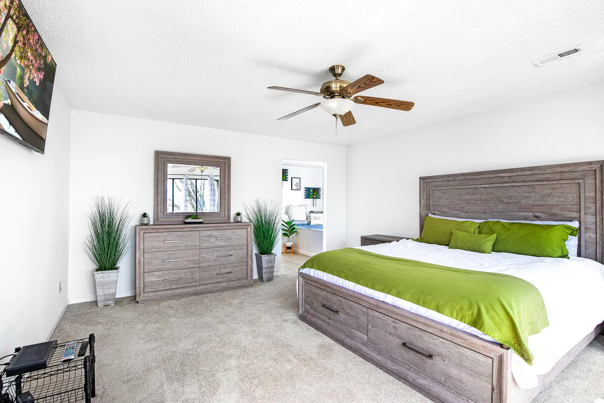 A serene bedroom with a cozy bed adorned with green accents, a decorative mirror above the headboard, and a ceiling fan. The room is bathed in gentle natural light, creating a restful environment ideal for unwinding after a busy day.