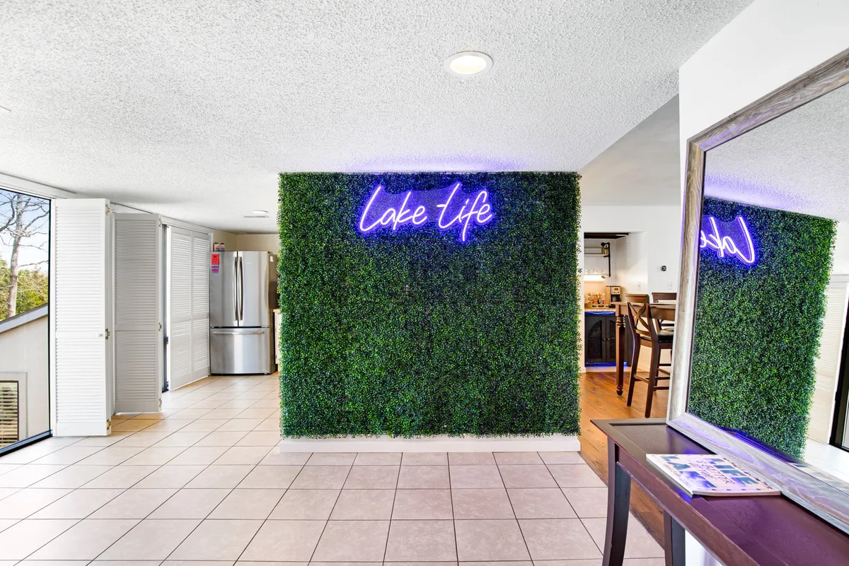 A bright, open space featuring a cheerful neon 'Lake Life' sign, lush greenery wall, and expansive windows that let in natural light and offer views outside. The layout is perfect for gatherings, meals, or simply relaxing in a vibrant communal area. The vibe is lively and connected to the outdoors.