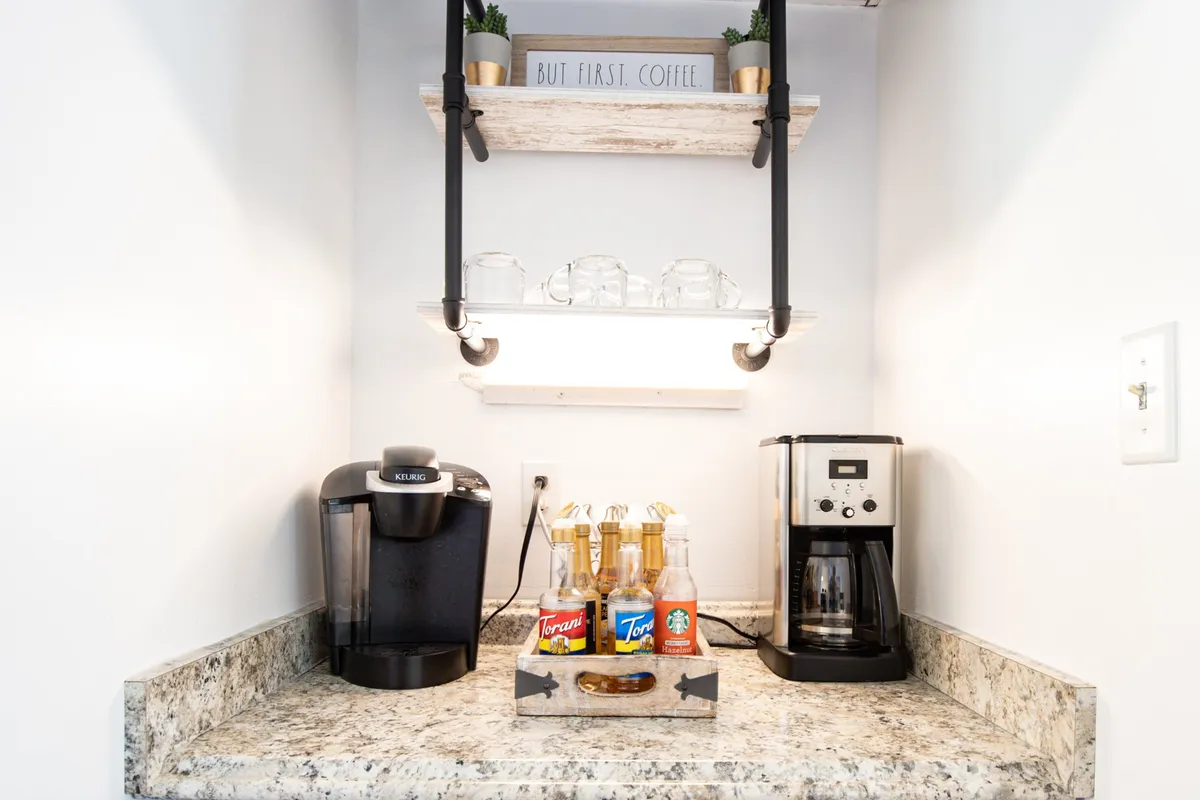 A cozy coffee bar setup with a coffee maker, bottled syrups, plant decor, and a warm wooden sign, inviting guests to create their favorite brews and enjoy casual conversation.