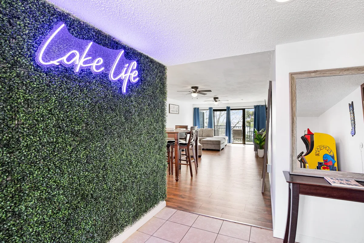 This image features a cheerful, open-plan living and dining area with a neon 'Lake Life' sign, green wall art, a dining table set for friends, and large windows offering sunset views. The space feels lively and social, perfect for group gatherings before or after a night out.