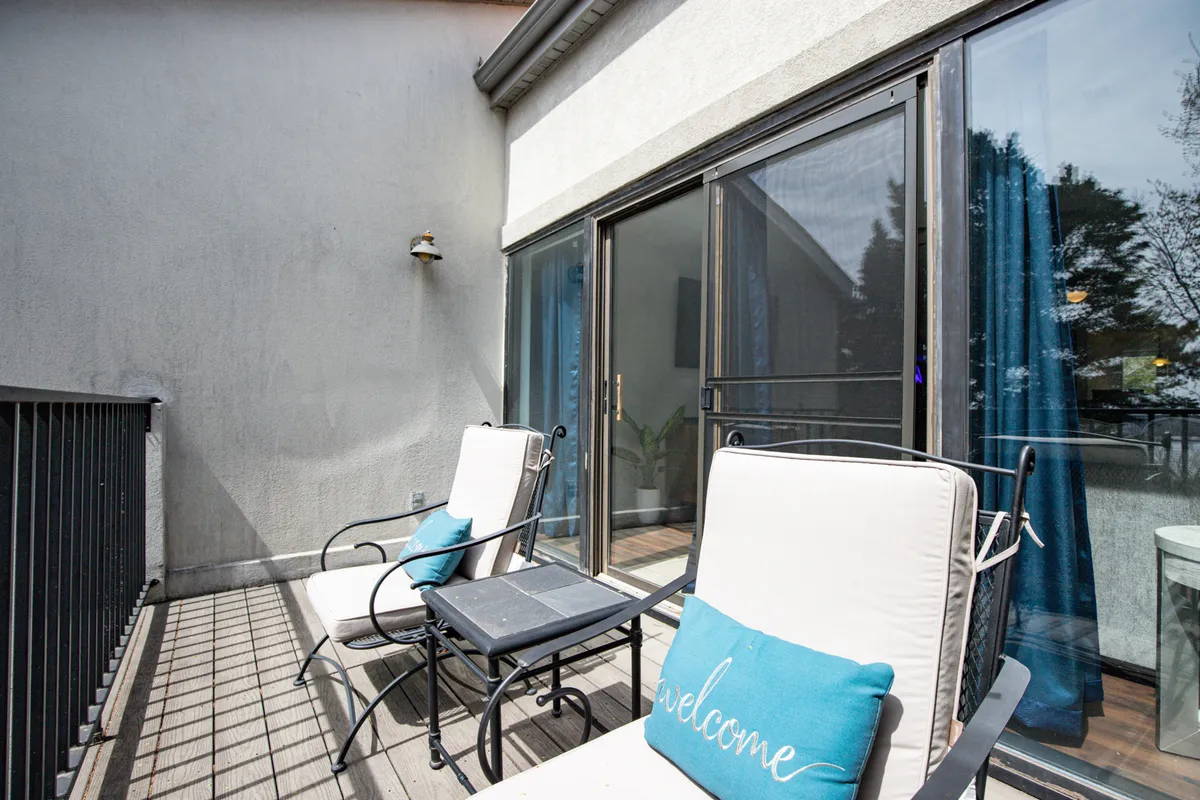 A cozy exterior seating area featuring two lounge chairs with a welcome pillow, large windows with curtains, and a relaxed atmosphere that suggests a perfect spot to relax with pets after a day exploring outdoors. The scene seamlessly connects the home to the surrounding environment, hinting at the lakeside vibe and outdoor adventure.