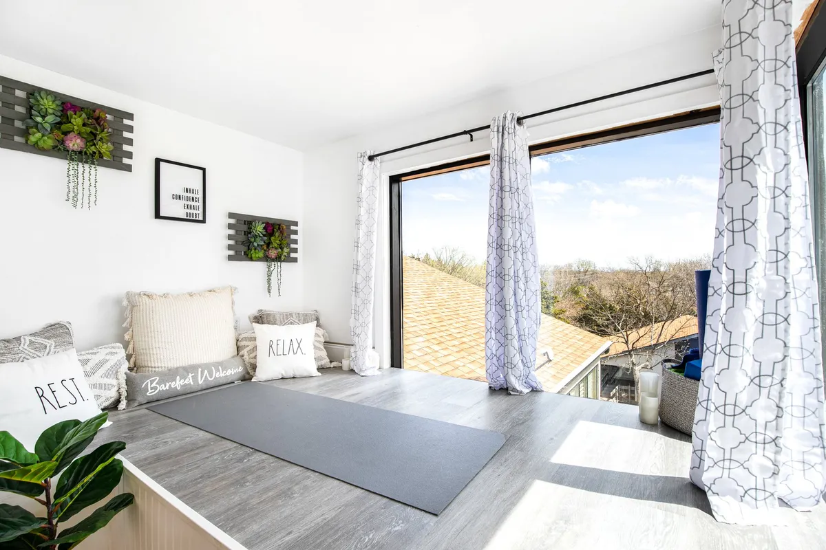 A tranquil yoga center space featuring a yoga mat positioned by a large window with views of the outdoors. Plush decorative pillows and lush indoor plants create a peaceful, inviting atmosphere ideal for mindful practices. The natural light streaming in enhances the serene, restorative mood.