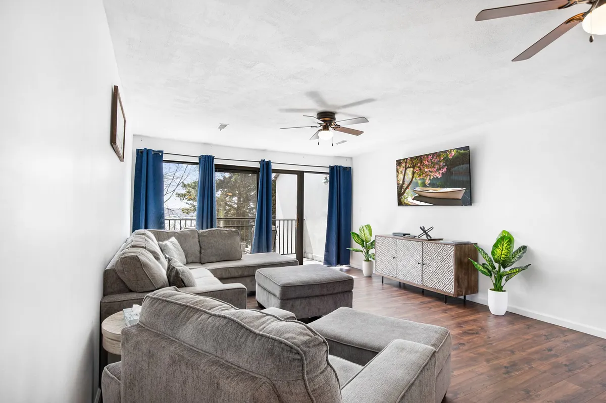 This image showcases a comfortable sectional sofa in the living area, a large TV with displayed artwork, an abundance of natural light from several windows, a decorative cabinet, and lush indoor plants. The space exudes a cozy, welcoming mood ideal for gatherings and relaxation.
