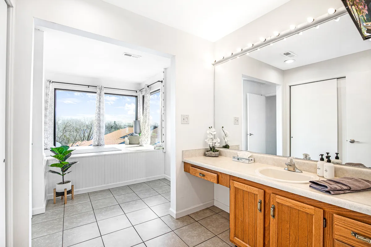 A modern bathroom featuring a spacious bathtub beneath a large window, a vanity with mirror, and a lush plant adding a touch of nature. Natural light fills the room, creating a spa-like atmosphere for self-care and relaxation.